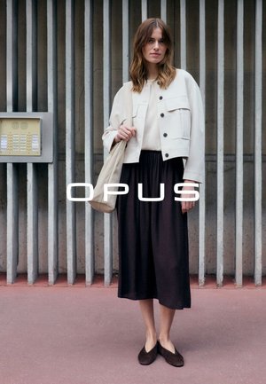 Femme portant une veste blanche, un haut crème, une jupe midi noire et des ballerines foncées, tenant un sac fourre-tout beige, debout devant un fond de barres métalliques verticales.