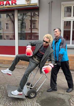 Person in einem grauen Anzug mit breitem Revers, roten Boxhandschuhen und Sonnenbrille sitzt auf einer Sackkarre. Eine andere Person in einer blauen Jacke steht dahinter.