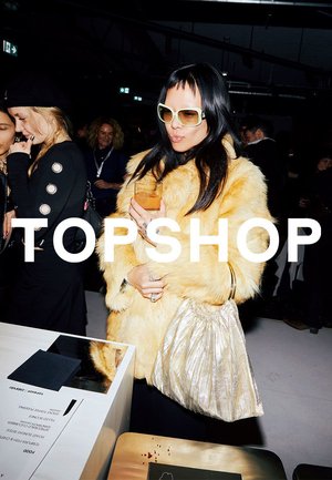 Donna con cappotto giallo in pelliccia sintetica e grandi occhiali da sole che tiene in mano una bevanda e una borsa metallica in un evento affollato al coperto con logo TOPSHOP.