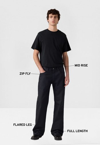 Vaqueros negros de talle medio con cierre de cremallera, pierna acampanada y diseño de longitud completa. Textura lisa de denim. Combinados con una camiseta negra sencilla.