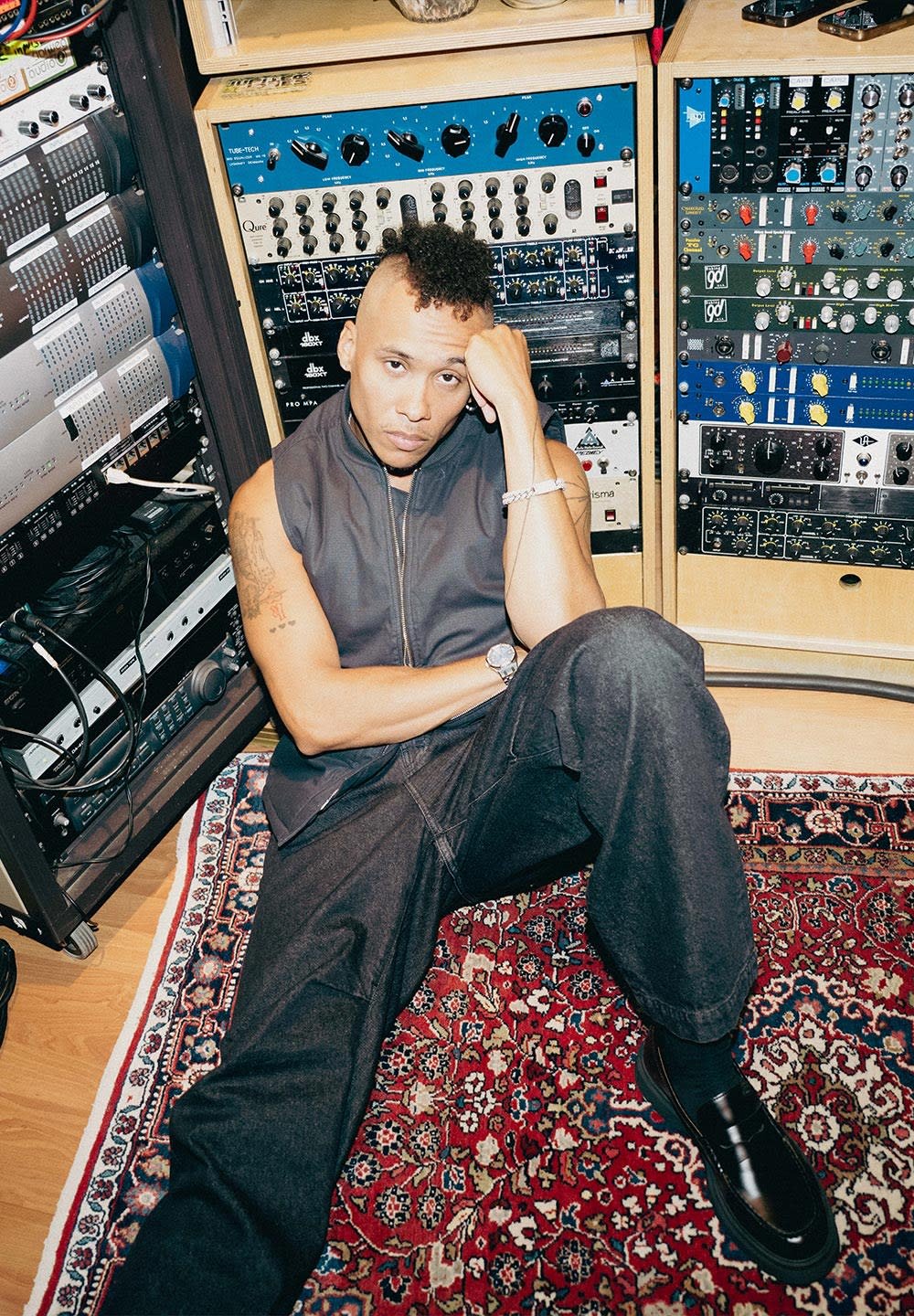 Person mit rasierten Seiten und lockiger Oberfrisur sitzt auf einem gemusterten Teppich und lehnt sich gegen die Audioequipment-Racks mit Knöpfen und Reglern im Tonstudio.