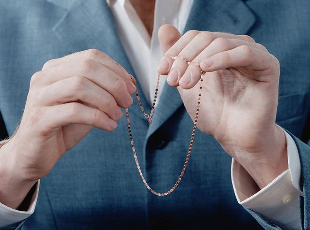 Αλυσίδα κολιέ σε ροζ χρυσό κρατημένη ανάμεσα σε δύο χέρια, με λεπτό, αλληλένδετο σχέδιο, λεία υφή και ασφαλές κούμπωμα.