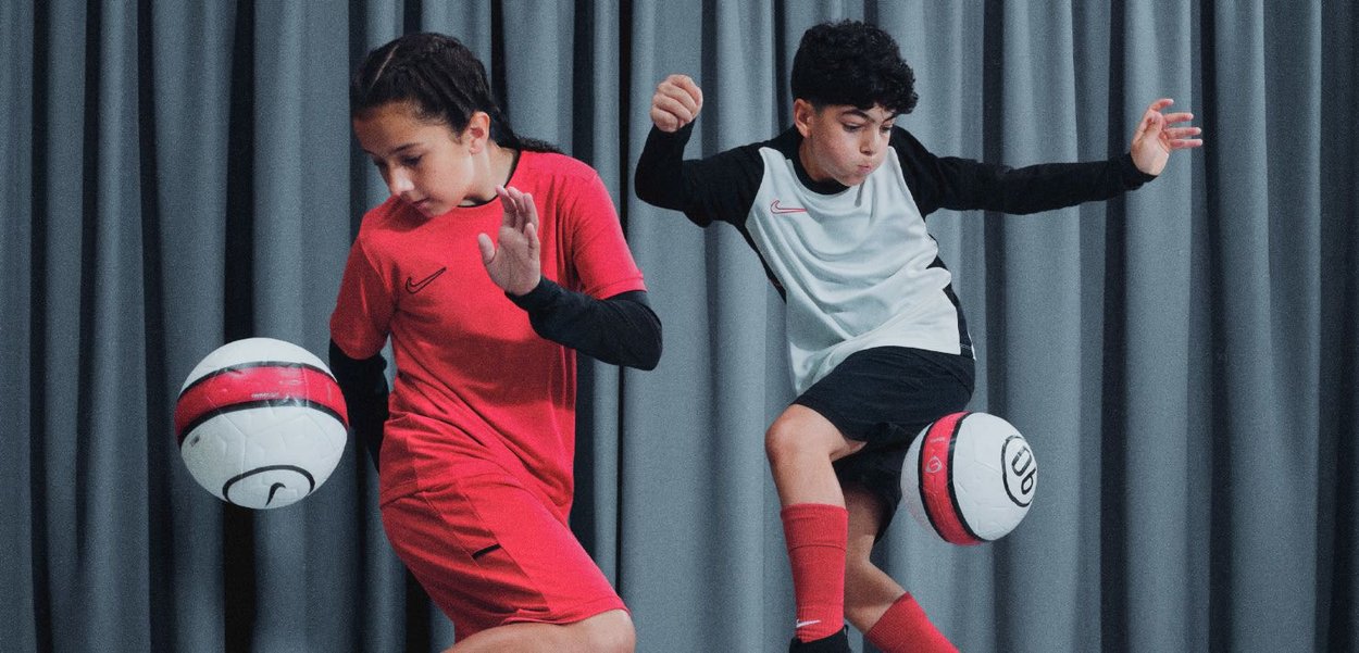 Deux enfants dribblent des ballons de football. L'un porte un maillot rouge et un pantalon noir ; l'autre a un maillot blanc et noir avec des accents rouges.
