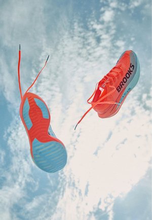 Bright orange running shoes with blue accents, lightweight mesh upper, thin laces, and distinctive outsole pattern, airborne against a blue sky.