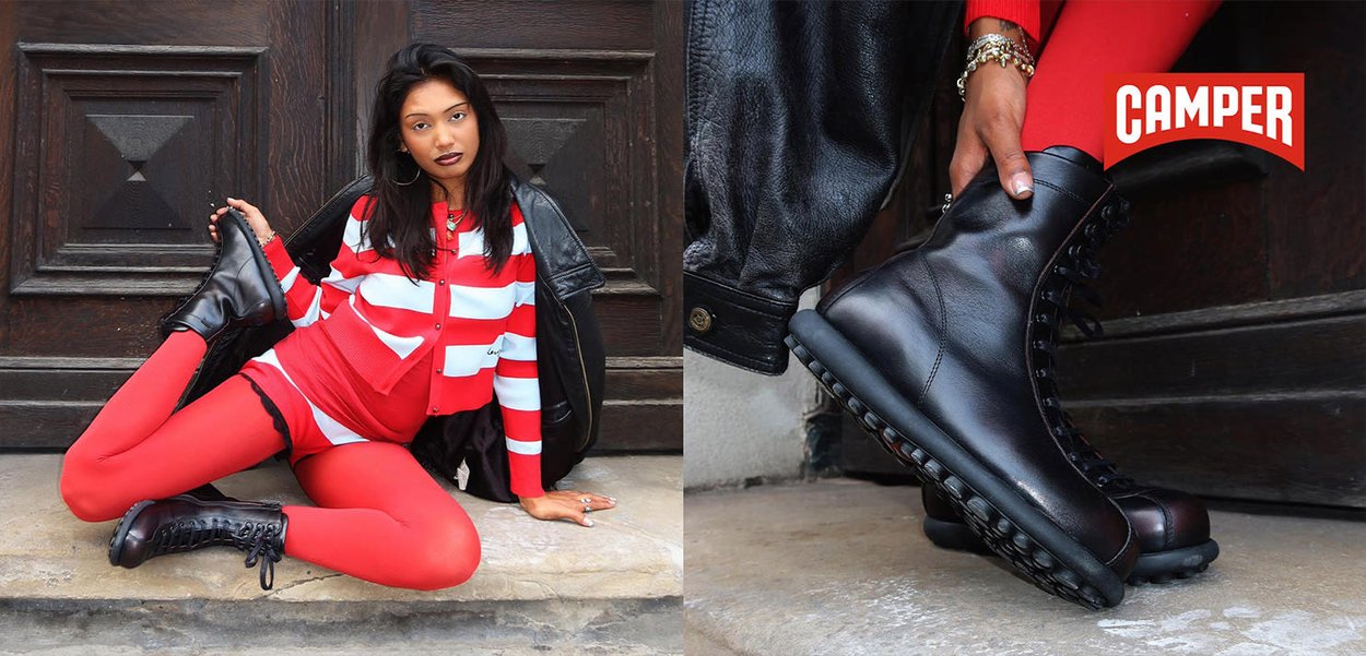 Woman in red and white outfit sitting by a wooden door, wearing black leather boots shown up close with Camper logo.