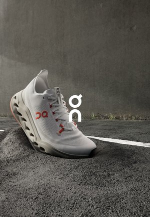Witte sportief schoen met een gestructureerde bovenkant, rode accenten en een kenmerkend chunky zoolontwerp. Gepositioneerd op een gestructureerd oppervlak.