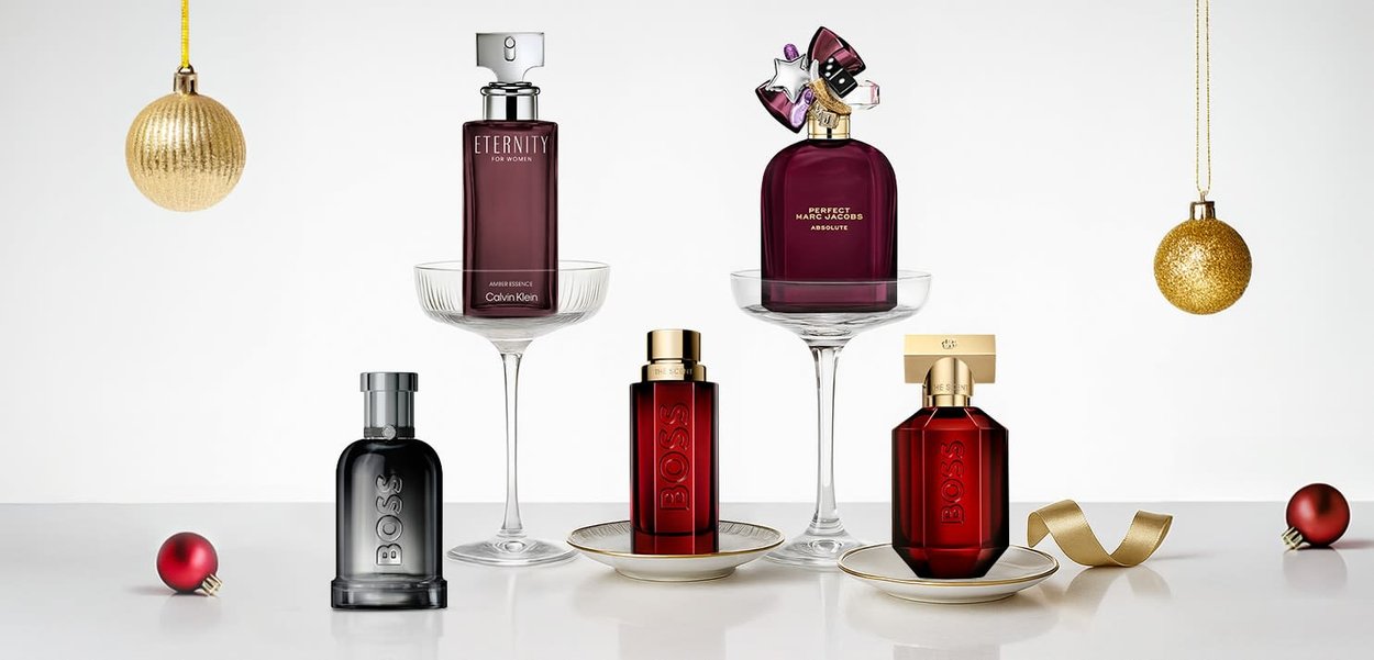 Cinque flaconi di profumo esposti: due in vetro scuro, uno con un tappo colorato e due in vetro rosso, su uno sfondo bianco con ornamenti.