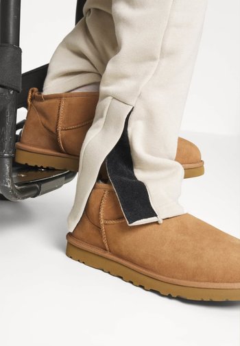 Ljusbruna mockakängor med platt sula, parat med beiga byxor som har en svart kardborreöppning längs sidokanten för lätt åtkomst.