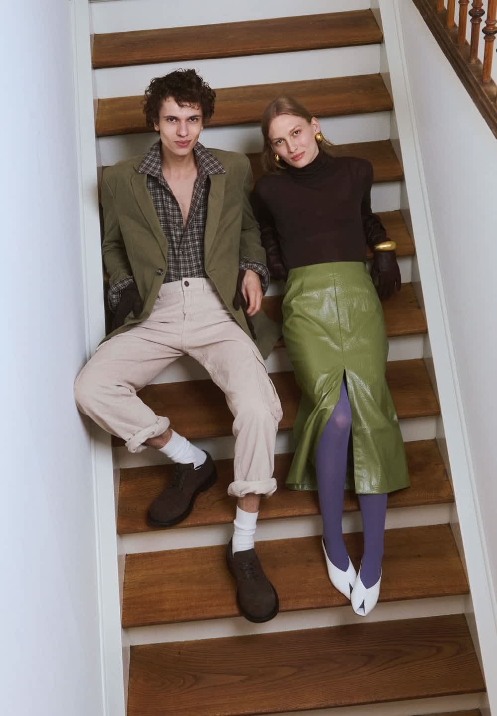 Två unga vuxna sitter på trappor av trä, iförda stiliga höstkläder med en grön läderskirt, lila strumpbyxor och en rutigt skjorta med en kavaj.