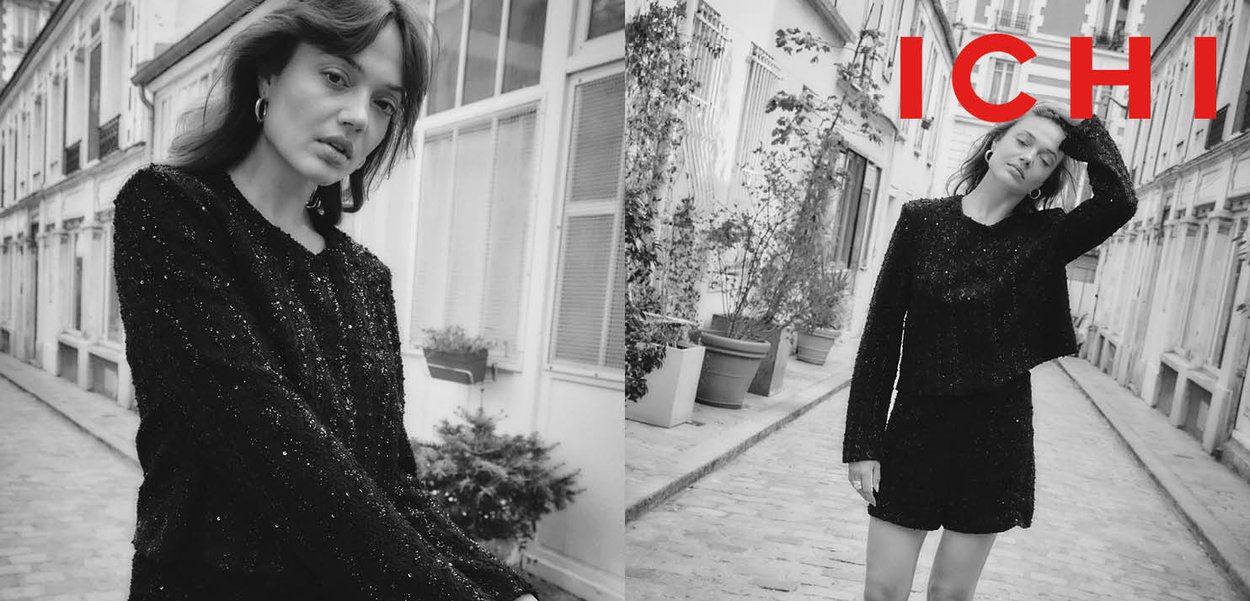 Black knitted sweater and shorts set with textured fabric, worn by a model. Background features a cobblestone street and potted plants.