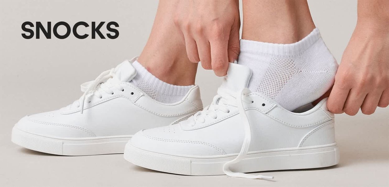 Hände ziehen weiße Knöchelsocken über Füße mit weißen Turnschuhen auf einem neutralen Hintergrund mit dem Wort SNOCKS.