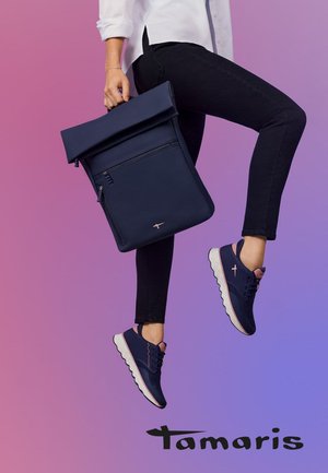 Sac à dos bleu marine à rabat, avec poche zippée, associé à des chaussures de sport bleu marine et rose, sur un fond dégradé rose.