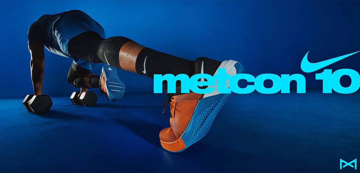Nike Metcon 10 sneaker in orange and blue, featuring a textured upper, padded collar, and a sturdy sole. Two dumbbells on a blue background.
