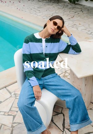 Grün-blau gestreifter Cardigan über einem weißen Oberteil, kombiniert mit hellblauen, weit geschnittenen Jeans. Model sitzt am Pool und trägt braune Sonnenbrillen.