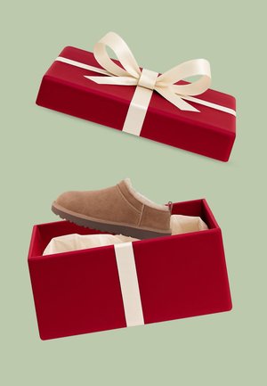 Brown slip-on shoe with fluffy lining displayed in a red gift box with a cream ribbon and bow, sitting on soft white tissue paper.