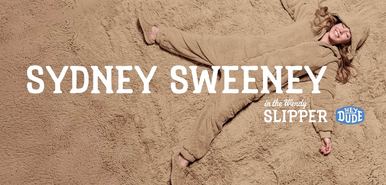 Beige, flauschiger, übergroßer Einteiler mit Kapuze, lange Ärmel und weicher Textur, vor einem passenden strukturierten Hintergrund.