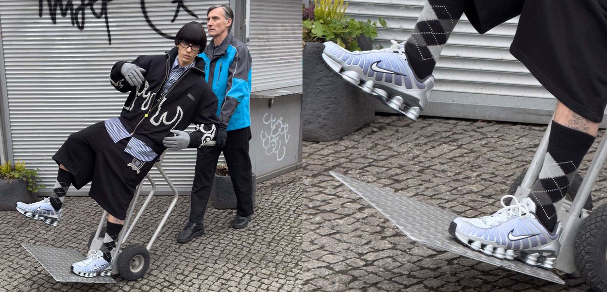 Svart jacka med dekorativa vita flammor, blå randig skjorta, svarta shorts, silvriga sneakers, handskar; person på en metallvagn med grå bakgrund.