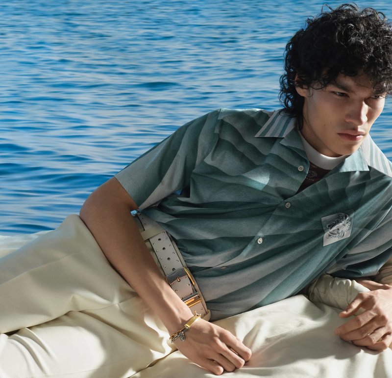 Jeune homme aux cheveux bouclés, allongé sur un bateau, portant une chemise verte à rayures, un pantalon beige et une ceinture argentée, avec de l'eau en arrière-plan.