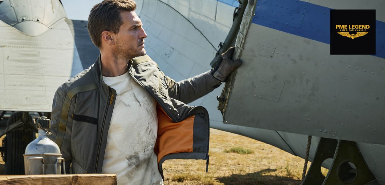 Man in leren jas en handschoenen inspecteert de staart van een vintage vliegtuig buiten onder een heldere hemel.