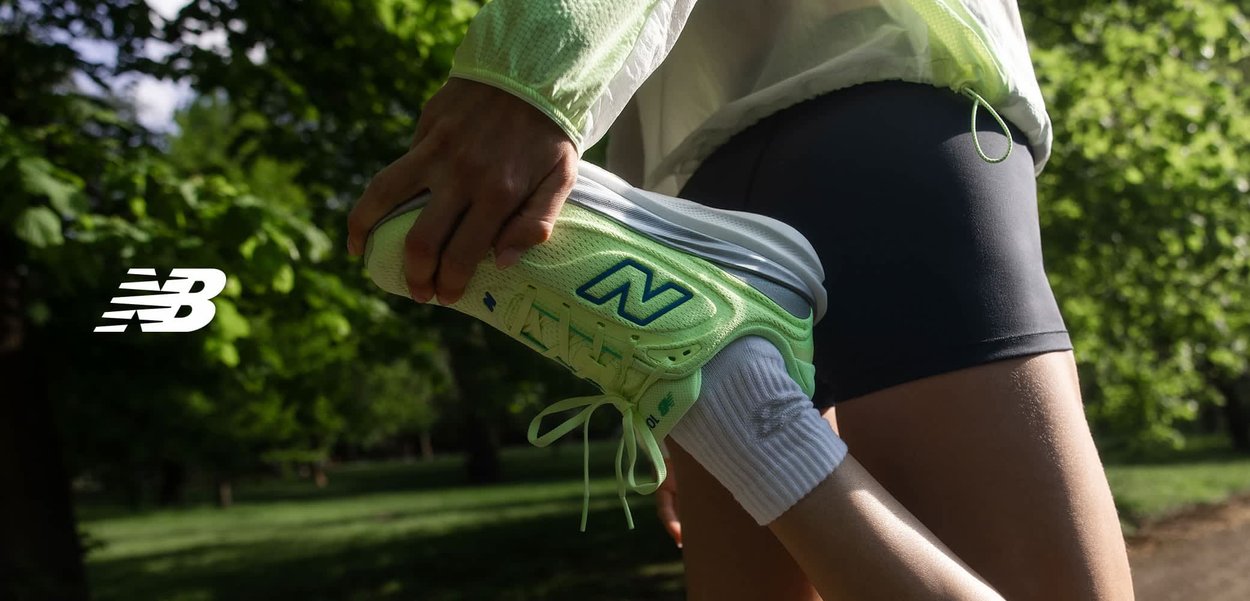 Coureur étirant la jambe, tenant une chaussure New Balance vert fluo, portant des shorts noirs et des chaussettes blanches, en plein air sur un sentier ensoleillé bordé d'arbres.