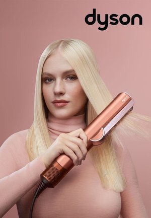 Donna con lunghi capelli biondi che utilizza una piastra per capelli Dyson color oro rosa su uno sfondo rosa con il logo Dyson sopra.