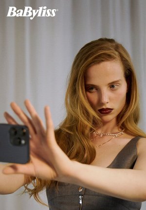 Modell med langt, bølgete blondt hår holder en smarttelefon, iført et grått plagg med dyp utringning, lagvis perlekjede og mørk leppestift.