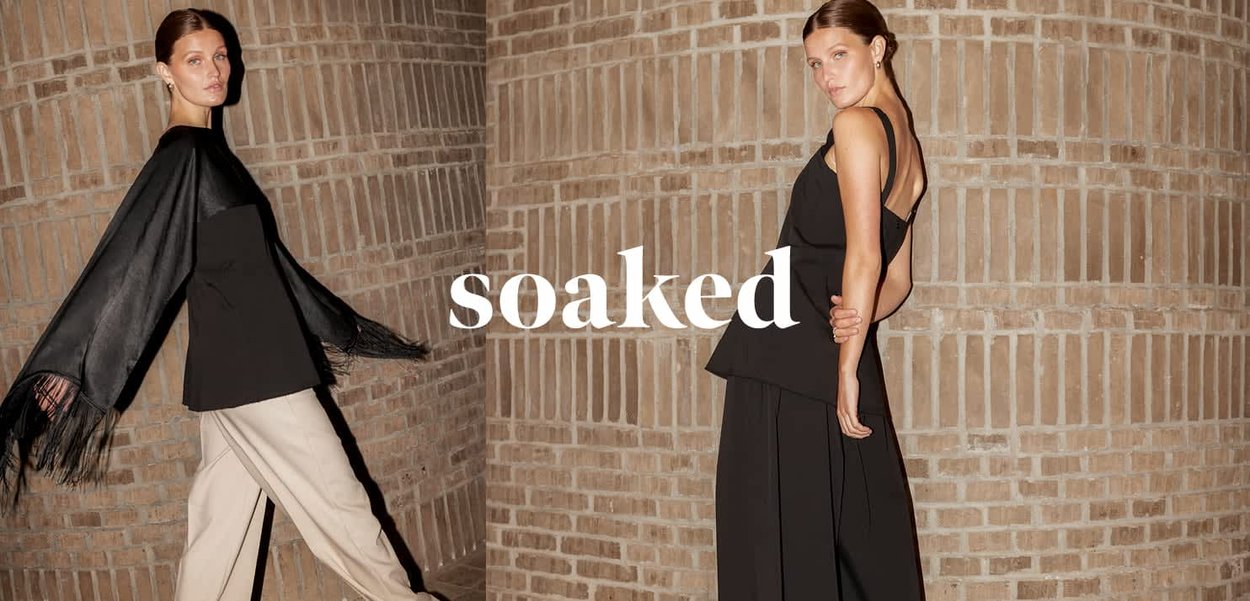 Zwei Frauen posieren vor einer Ziegelwand, tragen schwarze Oberteile; die eine trägt beigefarbene Hosen mit einem Fransenschal, die andere trägt schwarze, weit geschnittene Hosen.