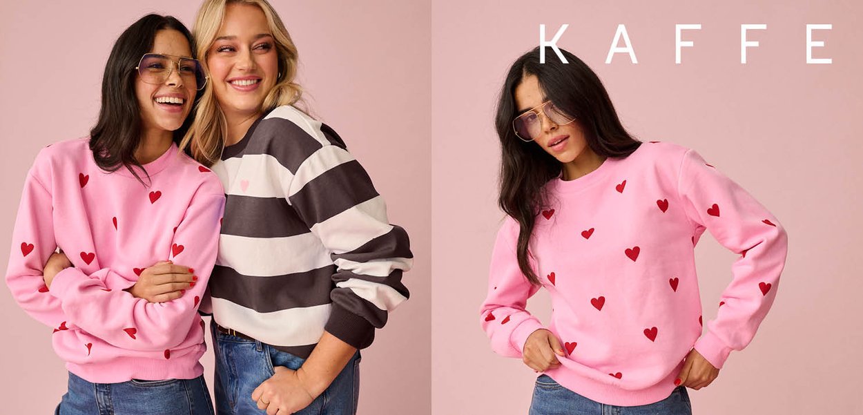 Zwei lächelnde Frauen; eine trägt ein rosa Sweatshirt mit roten Herzen und eine Brille, die andere ein schwarz-weiß gestreiftes Sweatshirt, beide in legeren Jeans.