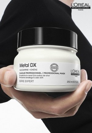 Hand som håller en burk med L'Oréal Professionnel Metal DX hårmask med svart lock, märkt för att förhindra brott och färgskiftning, 250 ml.