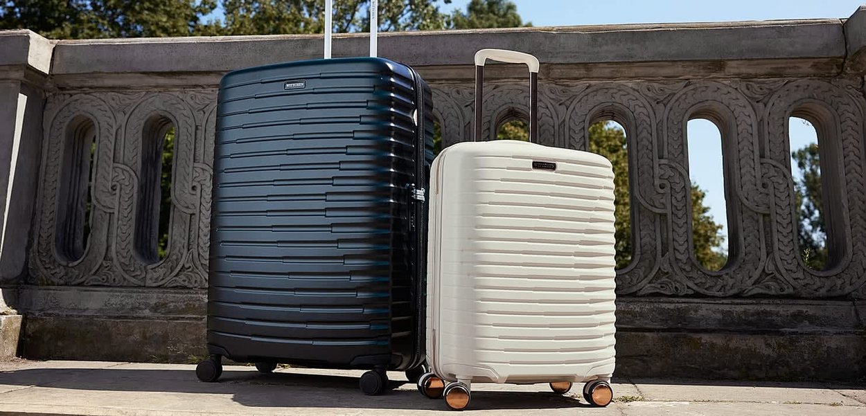 Zwei Hartschalenkoffer, einer schwarz und einer weiß, stehen auf einer Steinoberfläche mit einem dekorativen Steingeländer und Bäumen im Hintergrund.