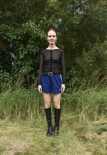 Schwarze transparente Langarmbluse mit Knöpfen, blaue Sportshorts, Nieten-Gürtel und kniehohe schwarze Stiefel. Das Model steht im hohen Gras.