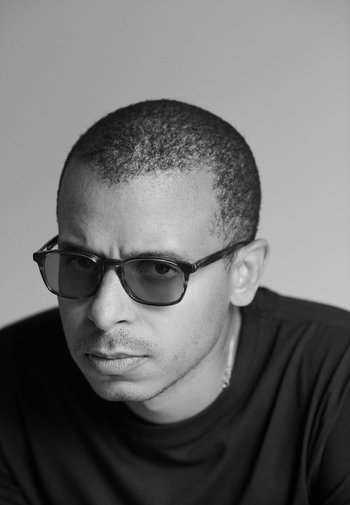Sort-hvidt billede af en person med kort, struktureret hår, iført mørke rektangulære briller og en sort t-shirt, der kigger direkte ind i kameraet.