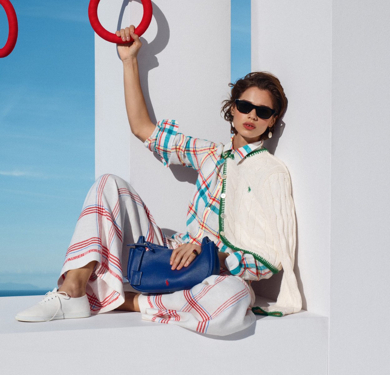 Mujer con gafas de sol, camisa de cuadros y pantalones anchos sentada contra una pared blanca, sosteniendo un bolso azul y agarrando un anillo rojo arriba.