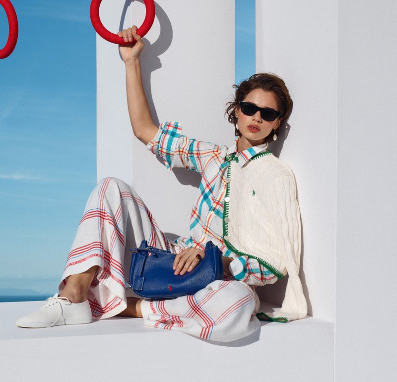 Mujer con gafas de sol, camisa de cuadros y pantalones anchos sentada contra una pared blanca, sosteniendo un bolso azul y agarrando un anillo rojo arriba.
