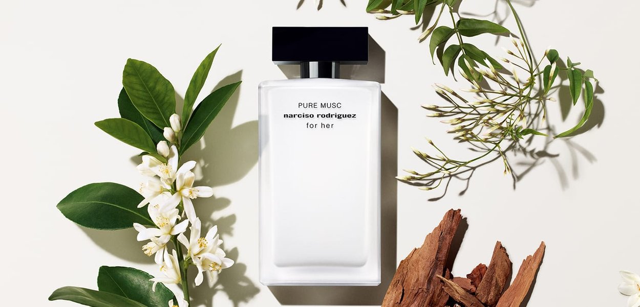 Clear rectangular glass bottle with a black cap, labeled "PURE MUSC narciso rodriguez for her," surrounded by green leaves, flowers, and brown wood pieces.