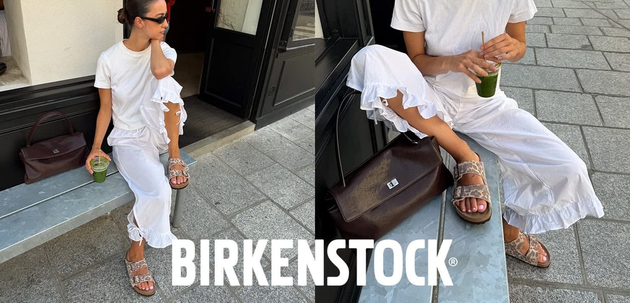 Frau in weißem, gerafftem Outfit und Sonnenbrille sitzt auf einer Steinbank, hält ein grünes Getränk und trägt Birkenstock-Sandalen mit einer braunen Ledertasche in der Nähe.