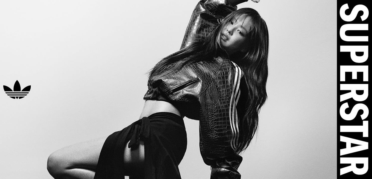 Donna posa in modo dinamico indossando una giacca e una gonna Adidas con tessuto texturizzato, con il logo Adidas a sinistra e la scritta "SUPERSTAR" a destra in lettere grassetto.