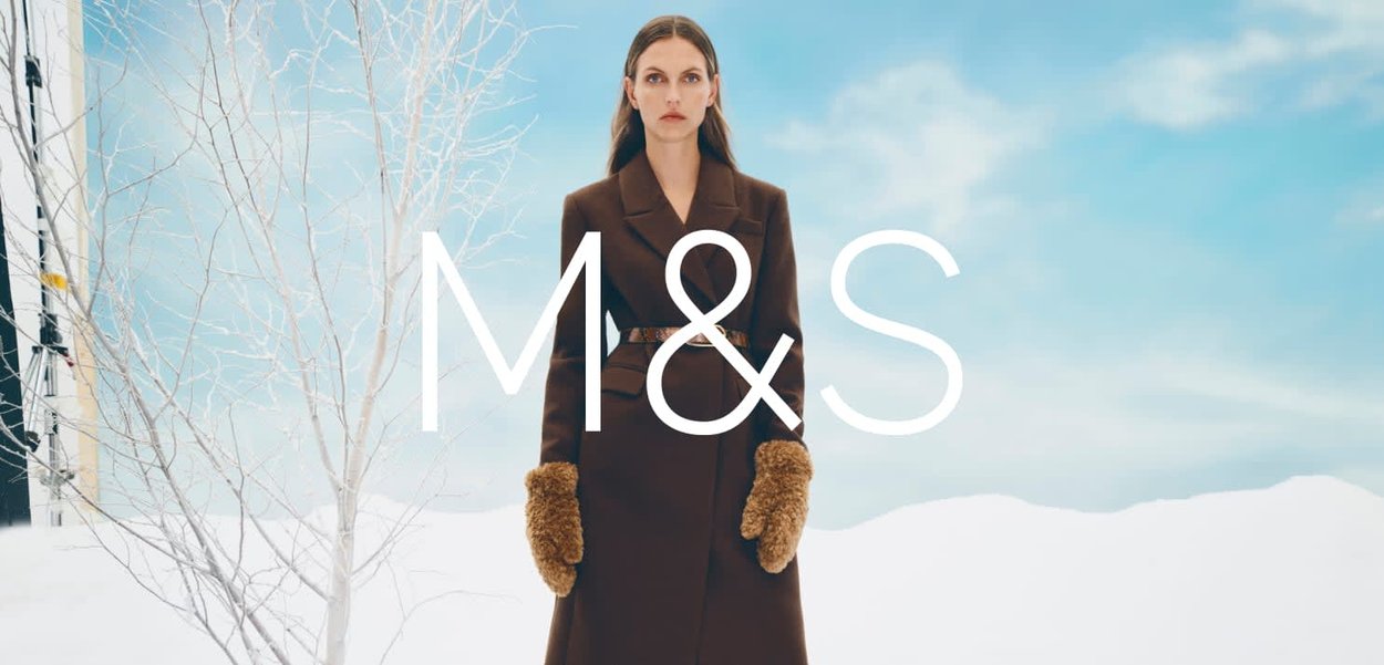 Brązowy wełniany płaszcz z paskiem, wyposażony w puszyste, jasnobrązowe rękawiczki. W tle jasnoniebieskie niebo i białe gałęzie drzew. Logo: M&S.