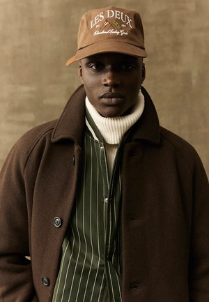 Brun uldfrakke over en grønstribet skjorte, parret med en hvid turtleneck og en brun kasket med "LES DEUX" broderet på forsiden.