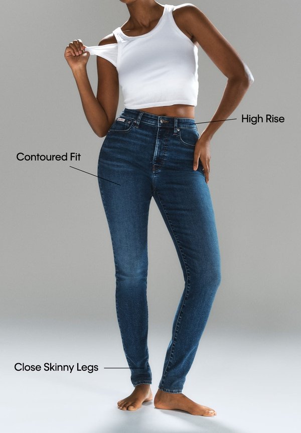 Jeans de tiro alto con un ajuste contorneado en denim azul oscuro. Piernas ceñidas al cuerpo. La modelo lleva una camiseta corta blanca y está descalza.