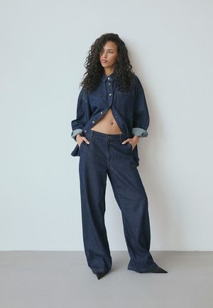 Jeansoutfit med en kort mörkblå skjorta med knäppning och högmidjade, vida byxor. Märkbara kontrasterande manschetter och fickor.