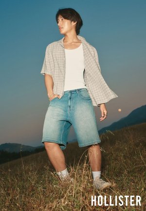 Een jonge man staat op een grasveld bij schemering, gekleed in denimshorts, een wit hemdje, een open kortärmlig overhemd, witte sokken en sneakers.