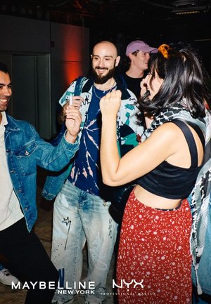 Quatre personnes souriantes et dansantes à l'intérieur lors d'un événement social décontracté, avec les logos de Maybelline et NYX en bas.