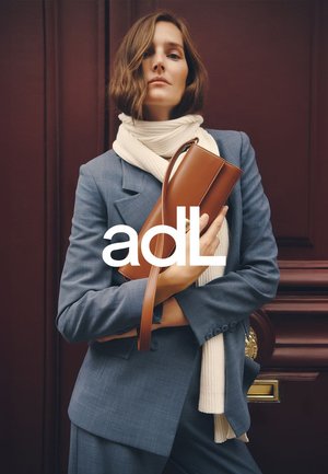 Sac en cuir marron avec une longue sangle, porté au bras d'un mannequin portant un costume bleu et une écharpe blanche. Design simple avec une texture lisse.