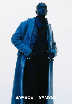 Lång blå kappa av mjukt material, lager-på-lager över en mörk skjorta och byxor. Har en halsduk och knappar, med synlig märkning nedtill.