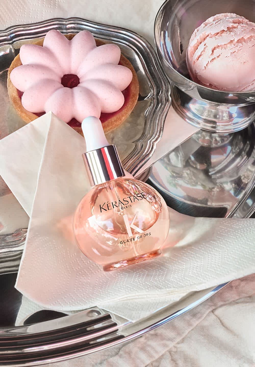 Kérastase Glaze Drops in a round glass bottle with a white dropper, set on a silver tray with a pink dessert and a bowl of ice cream.