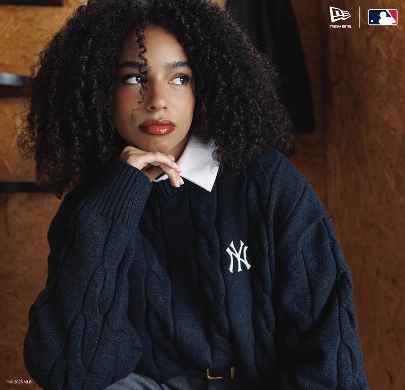 Donna con capelli ricci che indossa un maglione dei New York Yankees di colore blu navy, con il mento appoggiato sulla mano, che guarda di lato all'interno di un edificio.