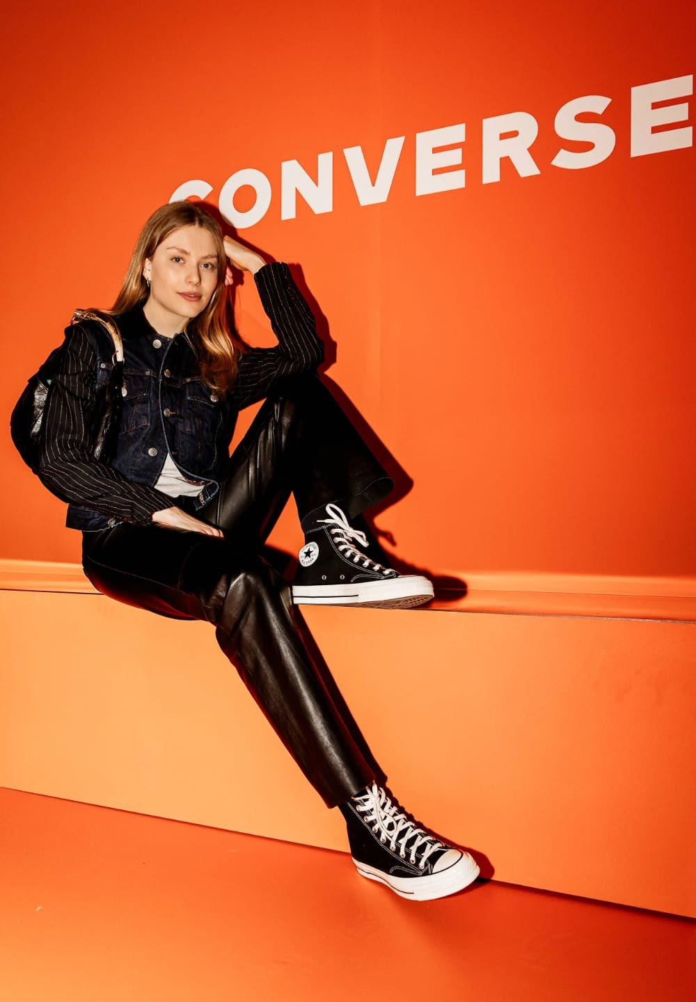 Black high-top Converse sneakers with white laces, paired with a denim jacket over a striped shirt and black leather pants against an orange background.
