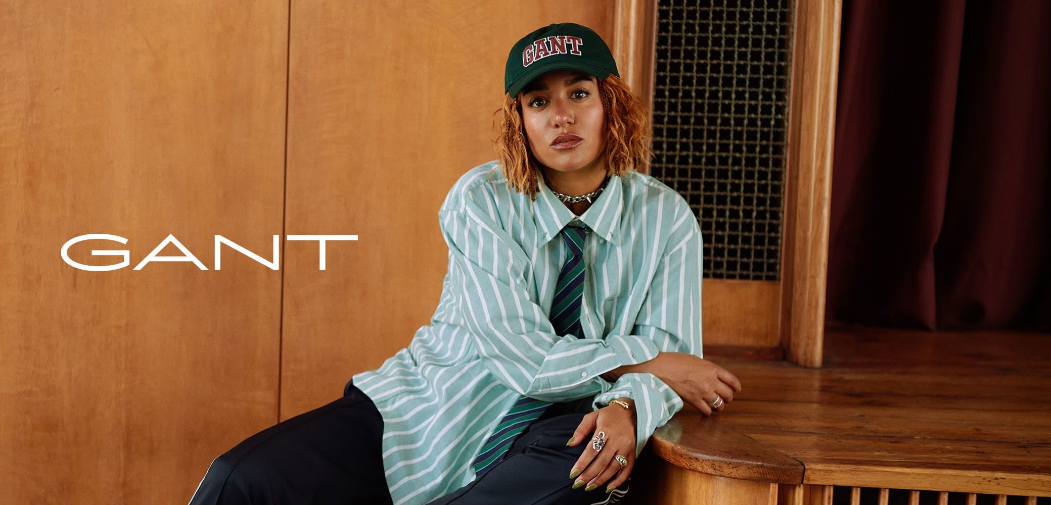 Green and white striped oversized shirt, black pants, green cap with red logo, silver accessories, seated against wooden background.