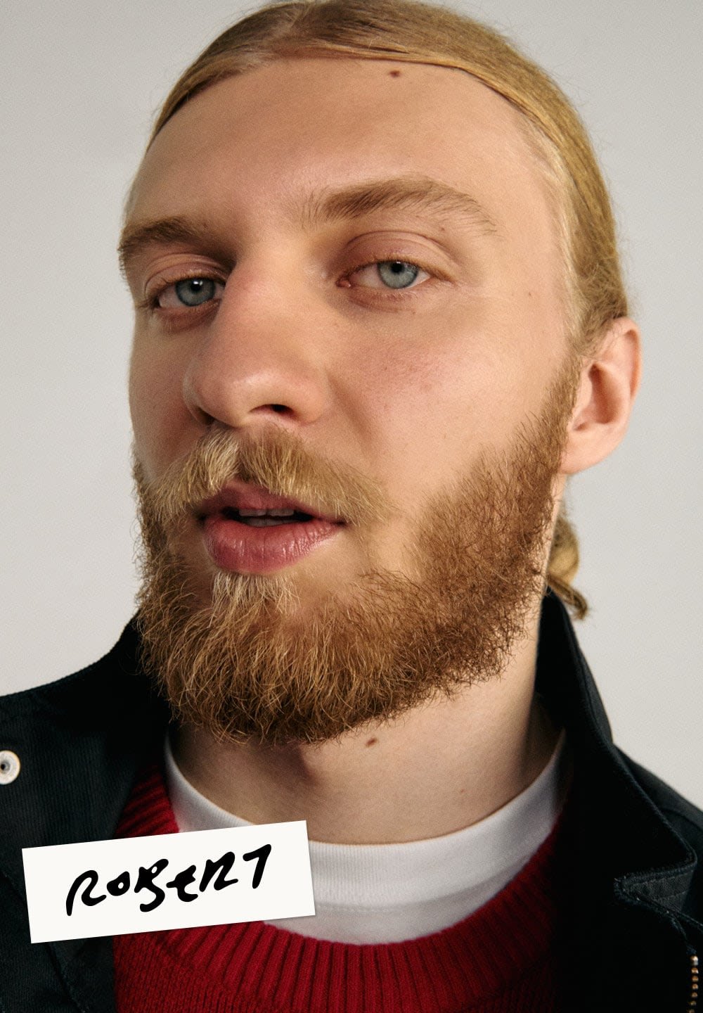 Portret mężczyzny z pełną brodą i długimi, jasnymi włosami, ubranym w czarną kurtkę na czerwony sweter, na neutralnym tle.